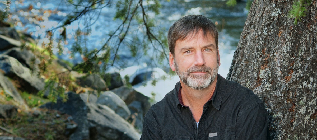 A man with pale skin, a greying beard, and a black button-down shirt stands on a riverbank leaning against a tree.